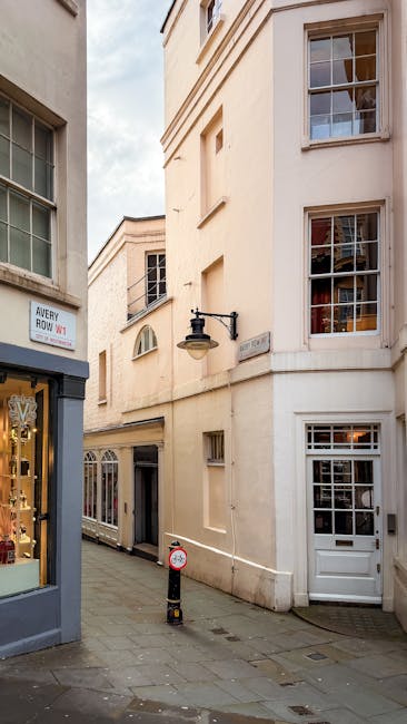 The facade of The Dorchester hotel in Mayfair features large glass doors with gold trim at the entrance, flanked by two men dressed in suits and face masks, possibly staff or security, standing on a p