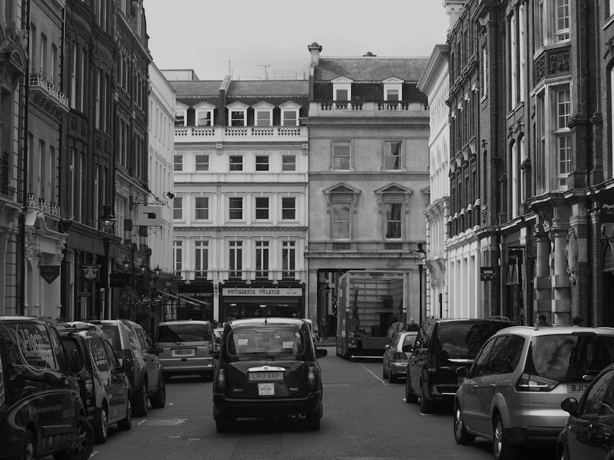 A street scene in an urban area shows a partially opened moving truck parked close to a historic multi-storey building with cream-colored stone facades and small sash windows. Several men are actively