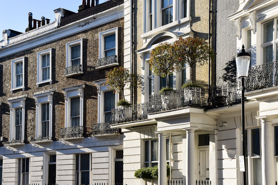 An overcast daytime scene of a quiet residential street in Mayfair, featuring wet pavement reflecting the surrounding buildings and sky. Classic red-brick and white-painted terraced houses with bay wi