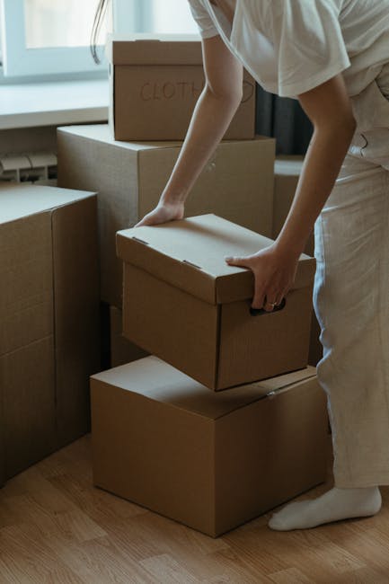 A man and a woman are inside a room surrounded by stacked cardboard boxes, some labeled with items such as 'books.' The man, wearing a grey hoodie, is kneeling on the floor and looking at the woman wi